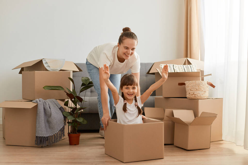 Portrét šťastné matky a dcery baví při stěhování v novém bytě, vyjadřující optimismus, holčička sedí v kartonové krabici se zvednutými pažemi, relokace, hraní si s matkou. - Fotografie, Obrázek