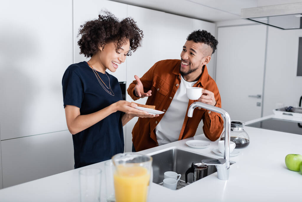 Mutlu Afrikalı Amerikalı adam mutfaktaki kız arkadaşının yanındaki sandviçi işaret ediyor.  - Fotoğraf, Görsel