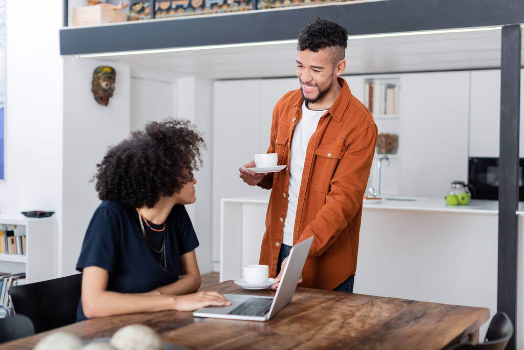 Mutlu erkek arkadaşının yanında dizüstü bilgisayar kullanan Afro-Amerikan kadının elinde kahve bardakları var. - Fotoğraf, Görsel