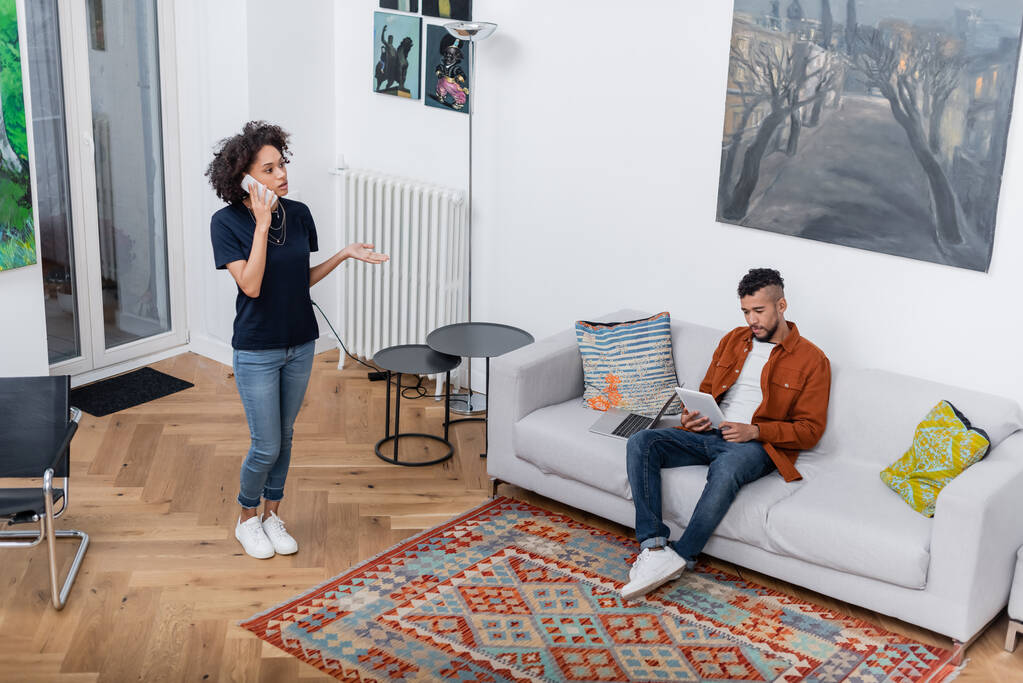 young african american woman talking on mobile phone while boyfriend using digital tablet in modern apartment  - Photo, Image
