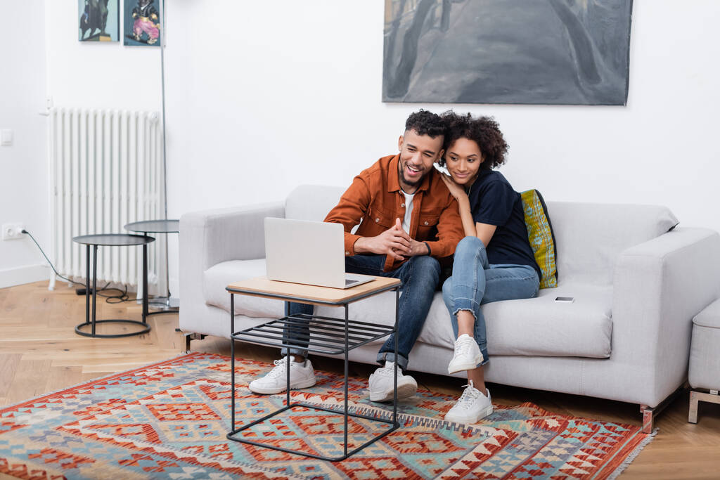joyeux jeune couple afro-américain souriant tout en regardant un film sur ordinateur portable dans un appartement moderne   - Photo, image