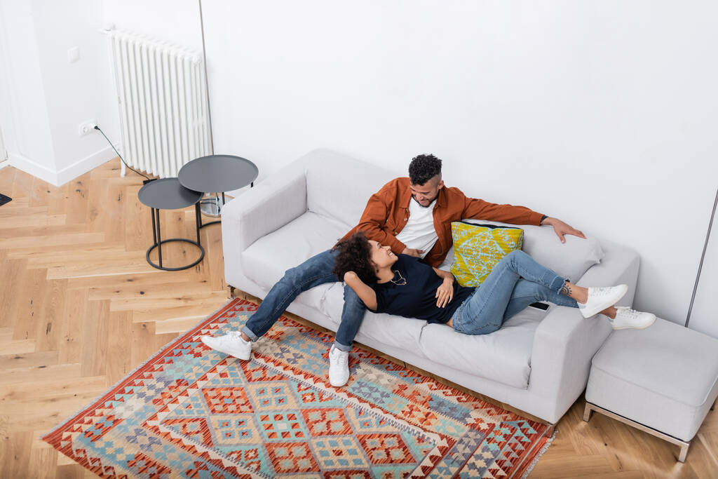 Modern apartmandaki kanepede dinlenirken birbirlerine bakan Afro-Amerikan çifti yüksek açılı görüyor.  - Fotoğraf, Görsel