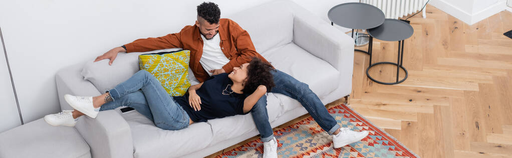 wysoki kąt widzenia wesoły afrykański para amerykańska patrząc na siebie podczas odpoczynku na kanapie w nowoczesnym apartamencie, baner - Zdjęcie, obraz