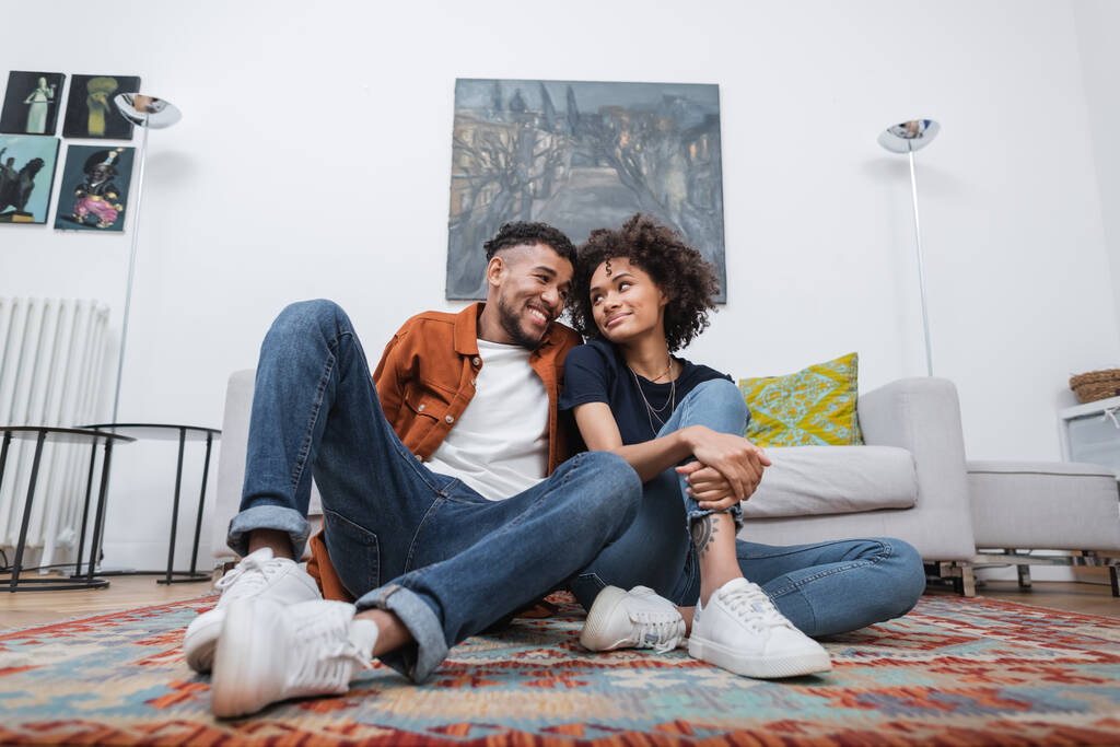 visão de baixo ângulo da mulher americana africana alegre com tatuagem e homem feliz olhando um para o outro enquanto sentado no tapete - Foto, Imagem