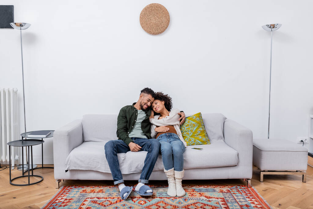 donna africana americana incinta seduta con marito felice sul divano in appartamento moderno  - Foto, immagini