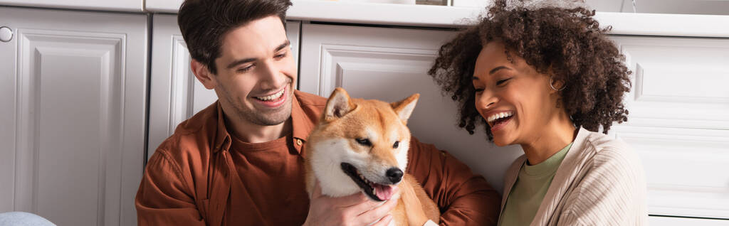 complacido interracial pareja sonriendo cerca divertido shiba inu perro en cocina, bandera - Foto, Imagen