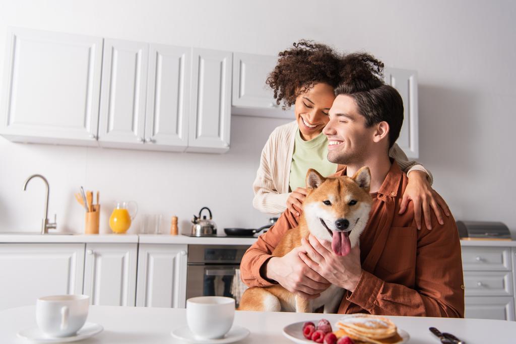 šťastný mezirasový pár usměvavý se zavřenýma očima v blízkosti shiba inu pes a snídaně v kuchyni - Fotografie, Obrázek