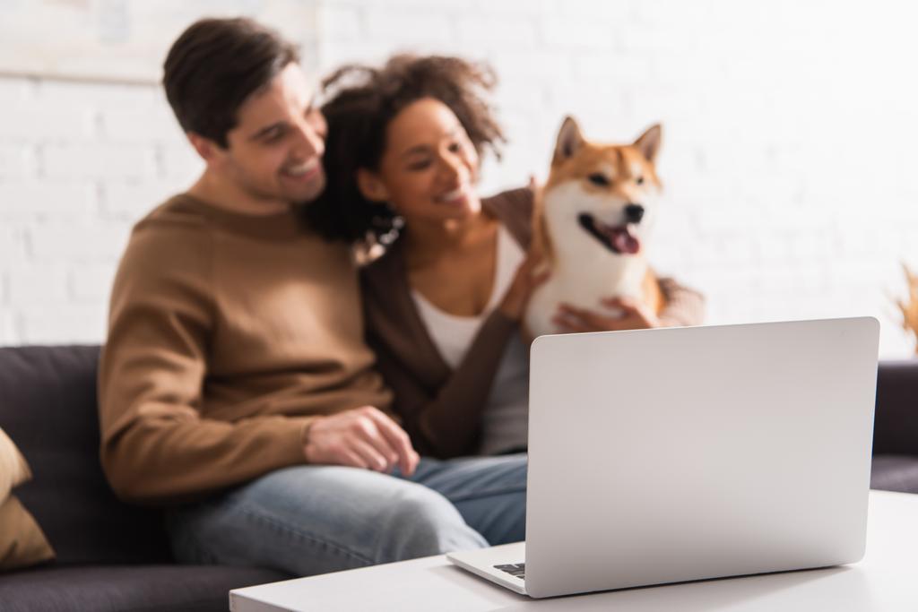 Evde Shiba Inu köpeği olan çok ırklı bir çiftin olduğu sehpanın yanındaki laptopta.  - Fotoğraf, Görsel