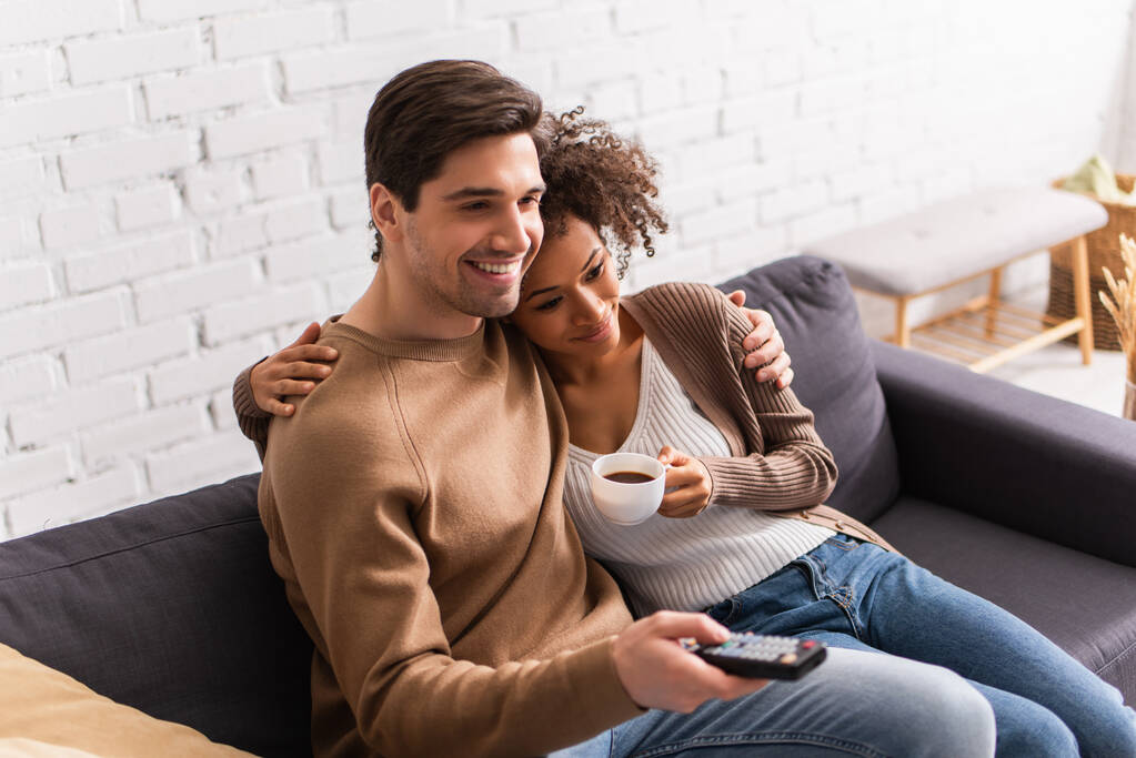 Sorridente donna afroamericana con caffè abbracciando fidanzato con telecomando a casa  - Foto, immagini