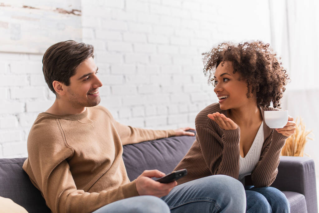 Sorrindo afro-americana com copo conversando com namorado com controle remoto em casa  - Foto, Imagem