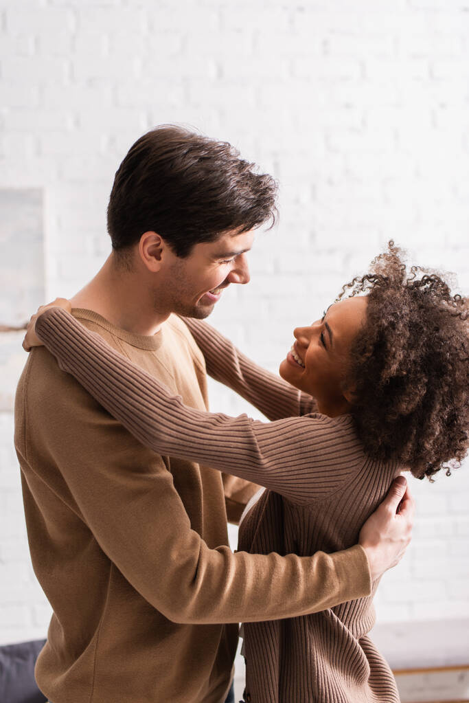 Cheerful interracial couple looking at each other while hugging at home  - Photo, Image