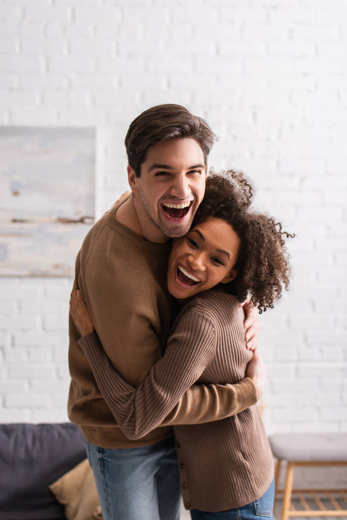 Positiva pareja multiétnica abrazando y mirando a la cámara en casa  - Foto, imagen