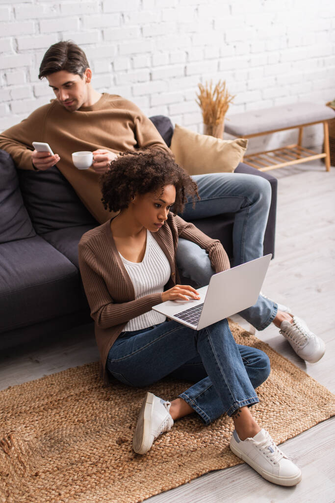 Libero professionista afroamericano che utilizza laptop vicino al fidanzato con caffè e smartphone sul divano di casa  - Foto, immagini