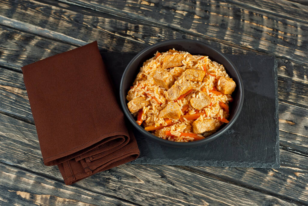 Pilaf with meat and carrots in a black bowl. Pilaf on a black background. Food on a shabby table. Bowl on an old wooden board. - Foto, Imagem