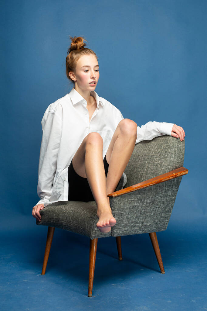Chica europea con el pelo rojo y la piel blanca aislado en azul sentado en la silla - Foto, Imagen