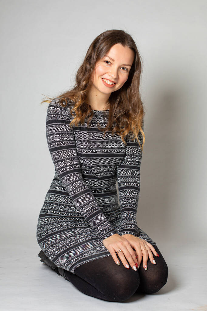 Lächelnde junge Frau in warmem gestreiftem Kleid mit langen lockigen Haaren, vereinzelt auf hellgrauen Knien sitzend - Foto, Bild