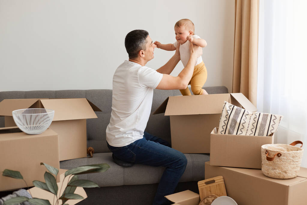 Портрет мужчины в белых футболках и джинсах, сидящего на диване с дочкой, распаковывающего коробки после переезда, играющего с ребенком, затаскивающего ребенка в картонную коробку. - Фото, изображение