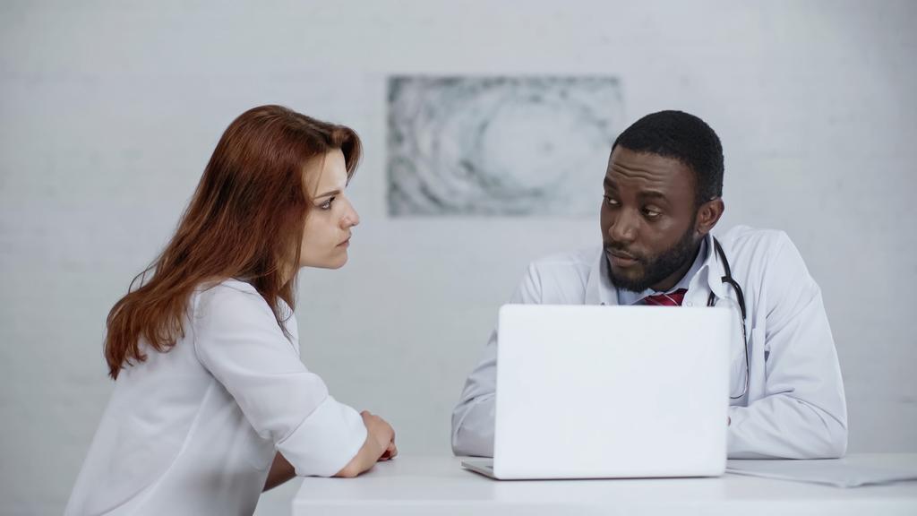 Afrykański amerykański lekarz rozmawiający z rudowłosym pacjentem przy laptopie na biurku - Zdjęcie, obraz