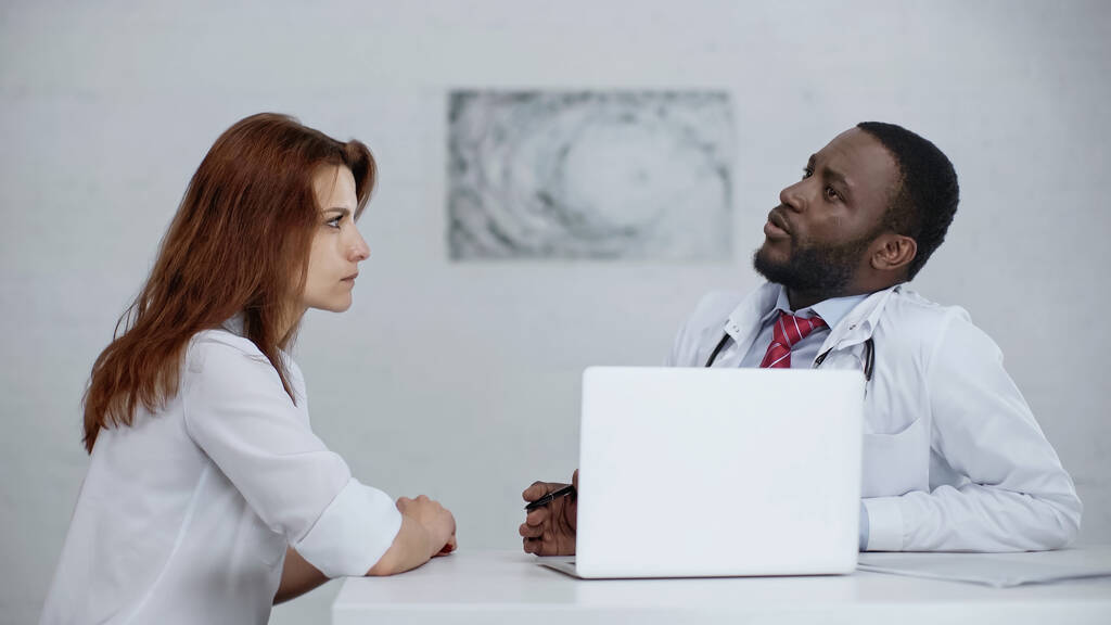 Afrykański amerykański lekarz rozmawia z rudą kobietą w pobliżu laptopa na biurku - Zdjęcie, obraz