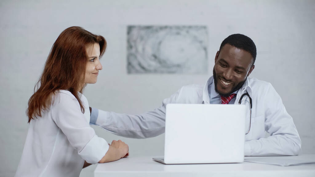 веселий афроамериканський лікар розмовляє з рудим пацієнтом біля ноутбука на столі
 - Фото, зображення