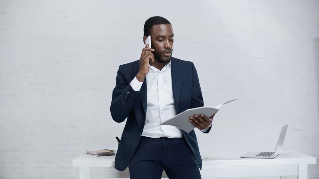 serious african american businessman talking on smartphone while standing with folder  - Photo, Image