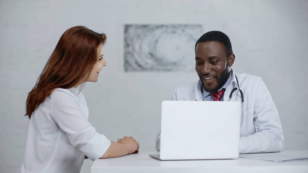 Mutlu Afrikalı Amerikalı doktor masadaki dizüstü bilgisayarın yanında kızıl saçlı hastayla konuşuyor.  - Fotoğraf, Görsel