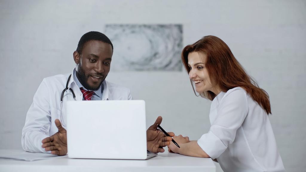 Afrikaans amerikaanse arts praten met vrolijke roodharige patiënt in de buurt laptop op tafel  - Foto, afbeelding