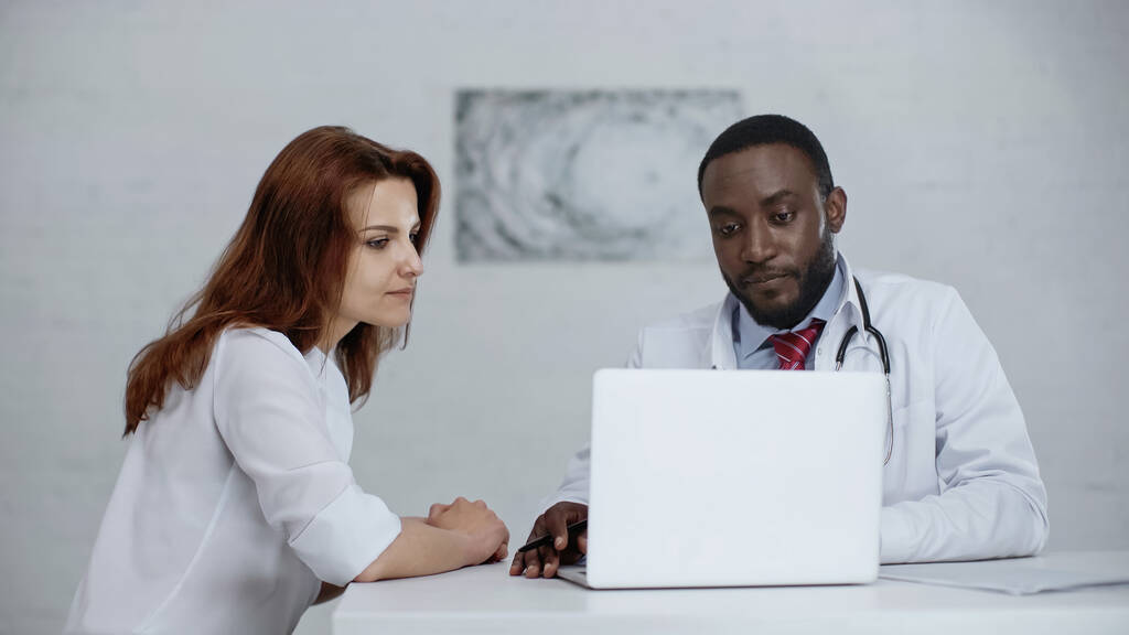 アフリカ系アメリカ人の医師と赤毛の患者がテーブルの上のノートパソコンを見て  - 写真・画像