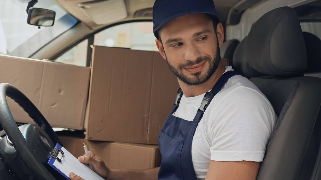 Smiling courier with clipboard sitting near cardboard boxes in auto  - Photo, Image