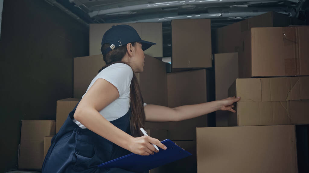 Mulher de entrega com prancheta de pé perto de caixas de papelão no carro  - Foto, Imagem