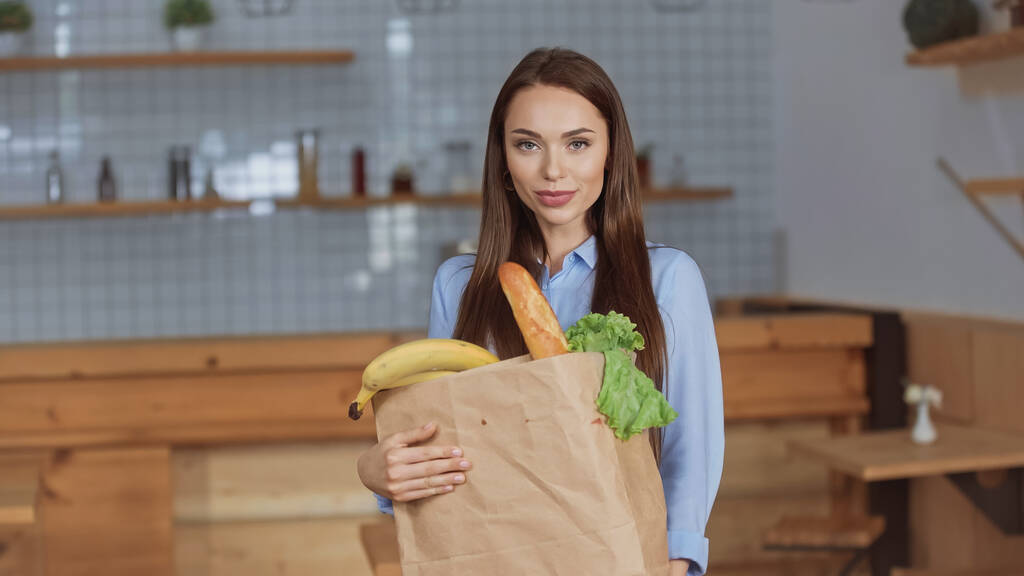 Csinos barna nő kezében csomag élelmiszer és nézi kamera otthon  - Fotó, kép
