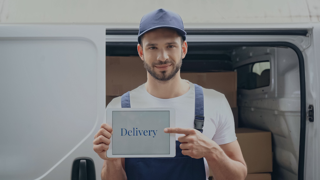 Χαμογελώντας delivery man δείχνοντας με ψηφιακή ταμπλέτα με γράμματα κοντά σε εξωτερικούς χώρους auto  - Φωτογραφία, εικόνα