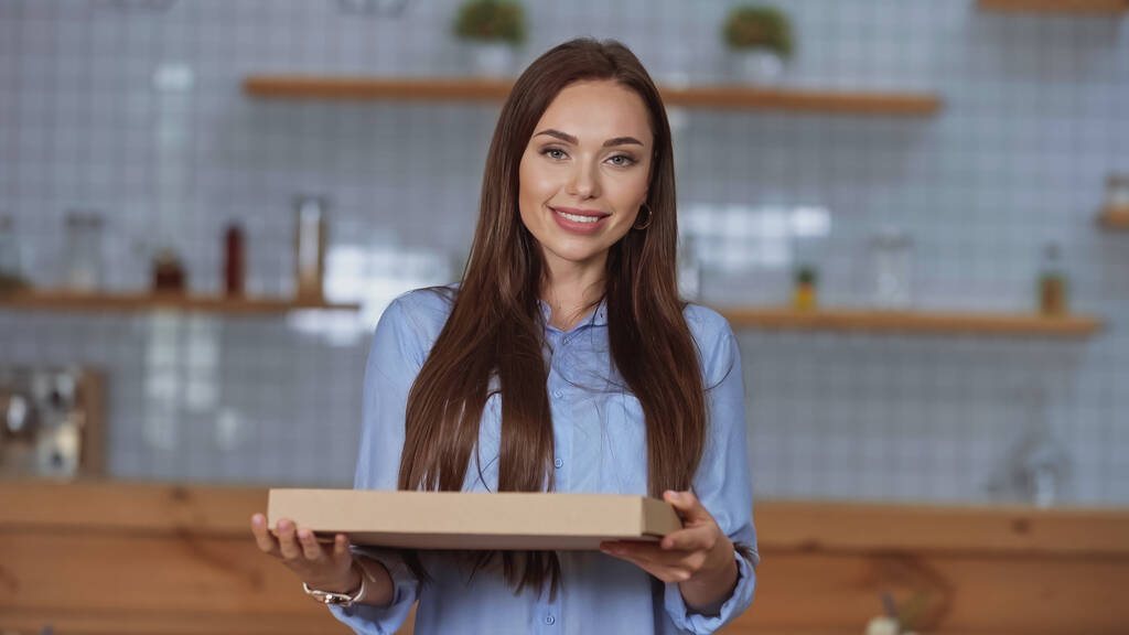 Mujer morena alegre sosteniendo la caja de pizza y mirando a la cámara en casa  - Foto, Imagen