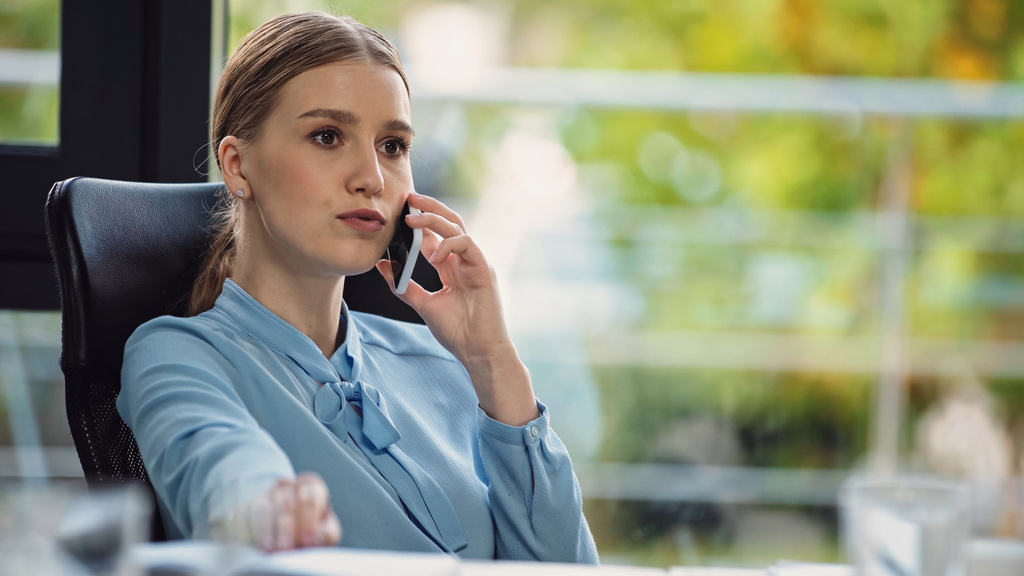 mujer de negocios pensativa llamando por teléfono celular mientras está sentada en el lugar de trabajo - Foto, Imagen