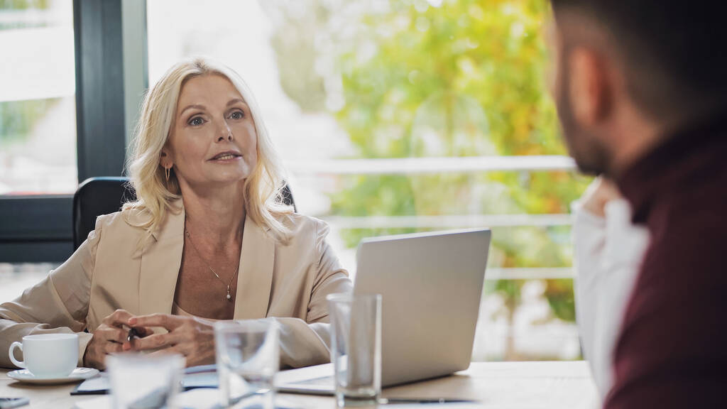 middle aged businesswoman talking to blurred colleagues near laptop in office - Photo, Image