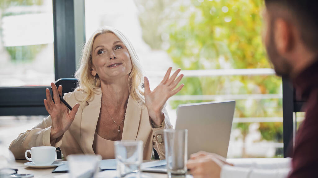 excited middle aged businesswoman gesturing near blurred colleagues in office - Photo, Image