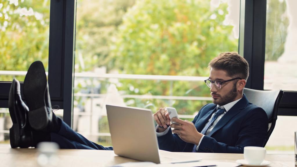 επιχειρηματίας με γυαλιά ηλίου κάθεται κοντά στο laptop με τα πόδια στο γραφείο και πληκτρολογώντας στο smartphone - Φωτογραφία, εικόνα