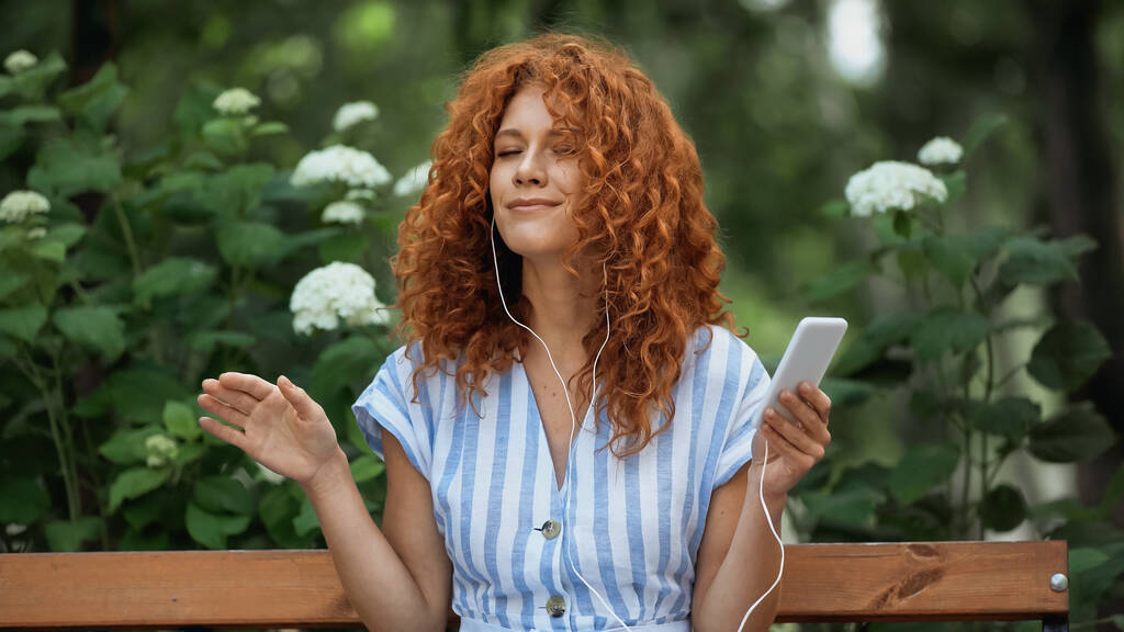 Neşeli kızıl saçlı kadın kulaklıkla müzik dinliyor ve parkta akıllı telefon kullanıyor.  - Fotoğraf, Görsel