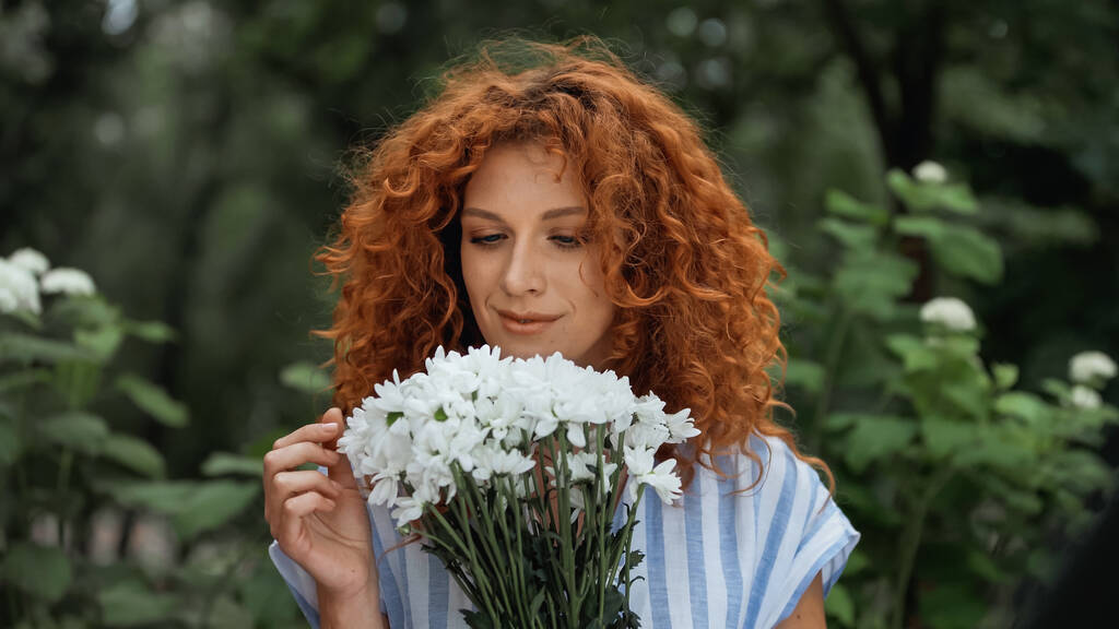 σγουρή κοκκινομάλλα γυναίκα κοιτάζοντας μπουκέτο από λευκά λουλούδια  - Φωτογραφία, εικόνα