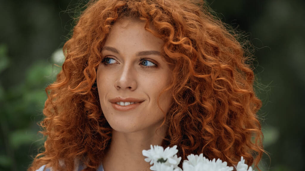 σγουρή κοκκινομάλλα γυναίκα με μπλε μάτια κοντά σε λευκά λουλούδια  - Φωτογραφία, εικόνα