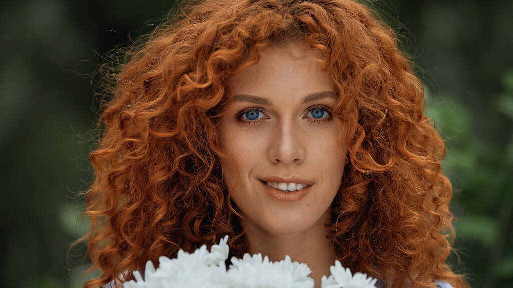 uśmiechnięta ruda kobieta z niebieskimi oczami w pobliżu białych kwiatów  - Zdjęcie, obraz