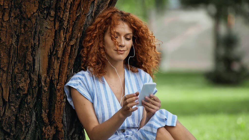 szczęśliwa ruda kobieta w niebieskiej sukience i słuchawki słuchanie muzyki podczas korzystania ze smartfona w pobliżu pnia drzewa - Zdjęcie, obraz