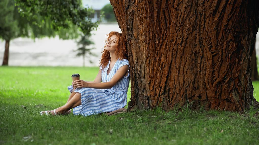 Uzun boylu mutlu kızıl saçlı kadın ağaç gövdesinin altında oturuyor ve elinde kağıt bardak tutuyor.  - Fotoğraf, Görsel