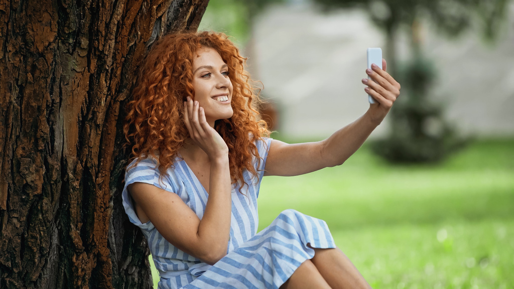 zadowolona ruda kobieta robi selfie siedząc pod pnia drzewa  - Zdjęcie, obraz