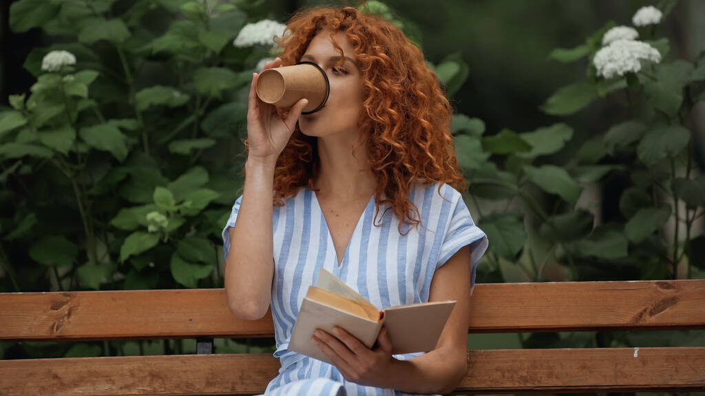 Çizgili elbiseli kıvırcık saçlı kadın kitap tutuyor ve parkta bankta otururken kahve içiyor.  - Fotoğraf, Görsel
