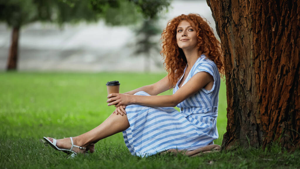 erfreut rothaarige Frau in blauem Kleid sitzt unter Baumstamm und hält Kaffee to go  - Foto, Bild