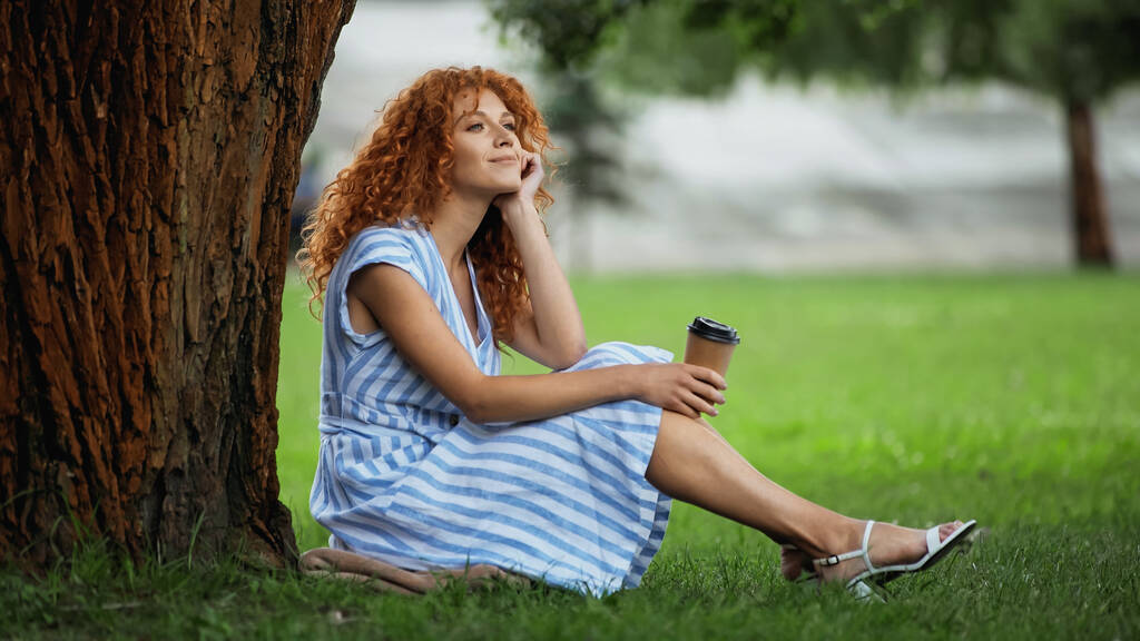 donna rossa sognante in abito blu sorridente mentre seduto sotto il tronco d'albero e tenendo il caffè per andare  - Foto, immagini