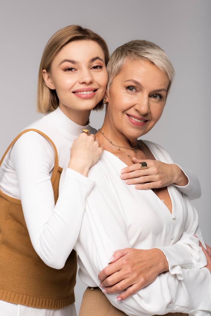 glücklich junge Tochter umarmt Mutter mittleren Alters und schaut in die Kamera isoliert auf grau - Foto, Bild