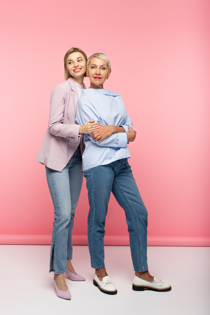 comprimento total de feliz jovem mulher abraçando mãe madura em jeans e blusa, enquanto em pé sobre rosa - Foto, Imagem