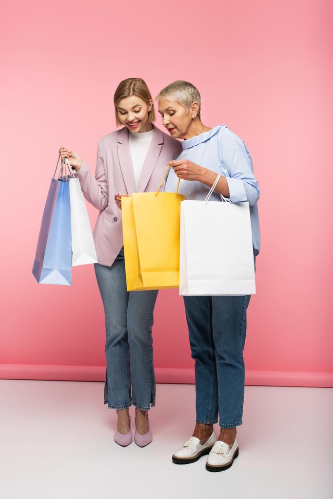 full length of happy woman and mature mother in jeans κοιτάζοντας τσάντες αγορών σε ροζ - Φωτογραφία, εικόνα